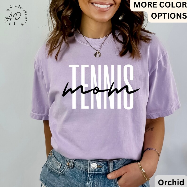 May include: A lavender t-shirt with the text "Tennis Mom" in white letters with black outlines. The text is in a stylized font with the word "Mom" written in cursive.