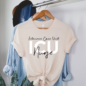May include: A cream-colored t-shirt with the text "Intensive Care Unit ICU Nurse" in black script and bold white letters. The shirt is hanging on a wooden hanger with a blue denim shirt and a brown hat in the background.