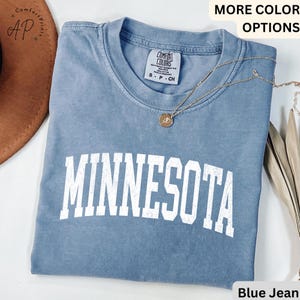 May include: A blue jean coloured t-shirt with the word "MINNESOTA" in white block letters. The shirt has a crew neck and short sleeves. A gold necklace with a pendant is draped across the shirt.