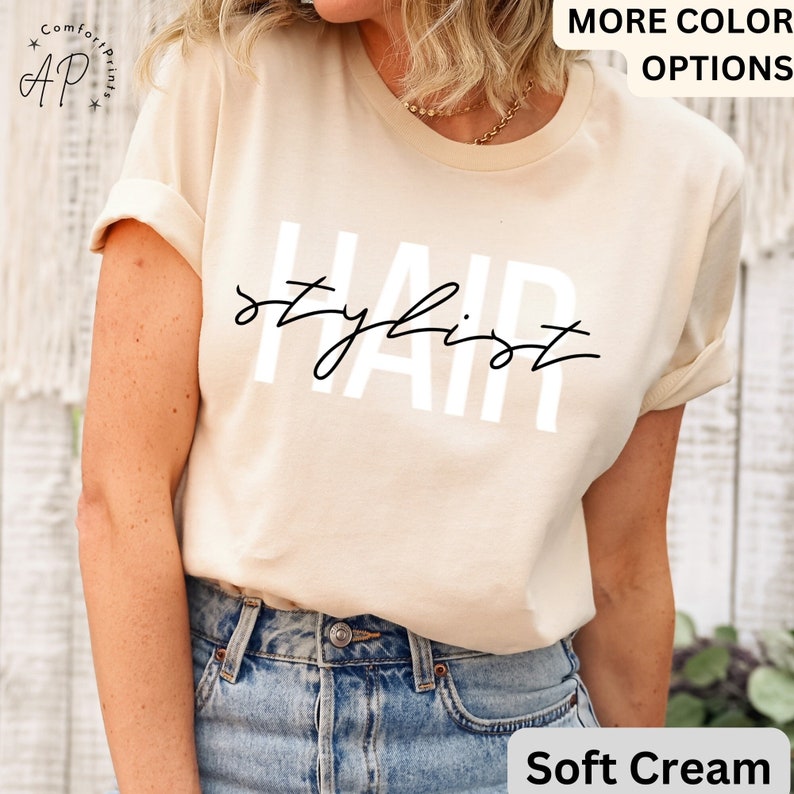 K&ouml;nnte beinhalten: Ein T-Shirt in Cremewei&szlig; mit dem Aufdruck "HAIR stylist" in Wei&szlig; auf der Vorderseite.