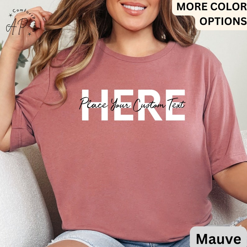 Puede incluir: Camiseta de color malva con la palabra "HERE" en letras may&uacute;sculas blancas grandes y el texto "Place Your Custom Text" en cursiva. La camiseta es de color liso y tiene mangas cortas. Las palabras "MORE COLOR OPTIONS" est&aacute;n en la esquina superior derecha.