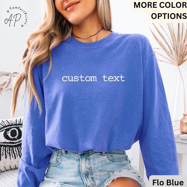 May include: A blue sweatshirt with the words "custom text" in white. The sweatshirt is a solid color and has long sleeves. The words "More Color Options" are in the top right corner. The sweatshirt is labeled "Flo Blue" at the bottom right.