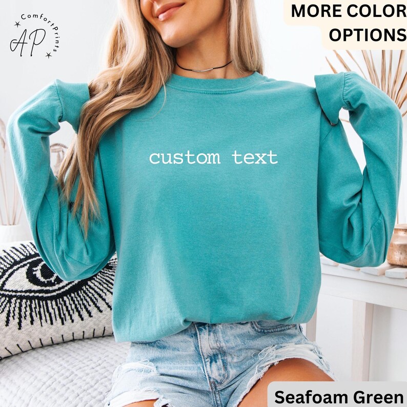 May include: Seafoam green long-sleeve sweatshirt with the text "custom text" in white. The sweatshirt has a crew neck and a relaxed fit. The image also includes the text "MORE COLOR OPTIONS" and "Seafoam Green".