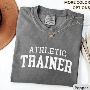 May include: A gray t-shirt with the words "ATHLETIC TRAINER" printed in white on the front. The shirt is made of a soft, comfortable material and has a vintage look.