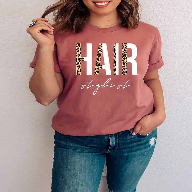 May include: A mauve t-shirt with the word "HAIR" in white letters with a leopard print pattern behind the letters. The text "stylist" is written in white cursive below the word "HAIR".