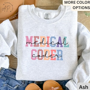 Peut inclure: Un sweat-shirt gris avec le texte "MEDICAL CODER" en couleurs arc-en-ciel. Le texte est en gras, avec une police de caractères en blocs.