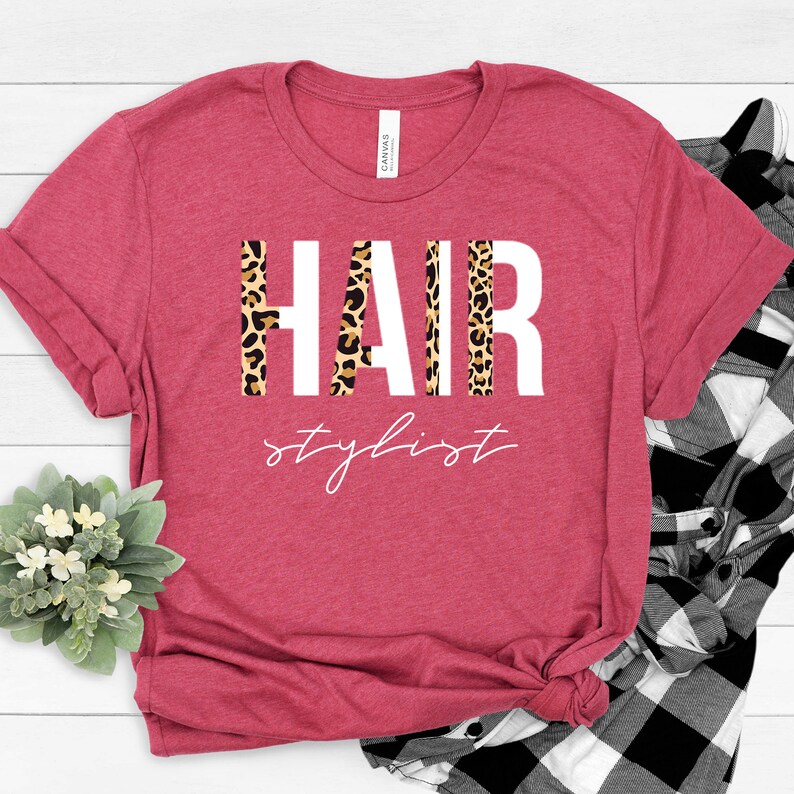 May include: A red t-shirt with the word "HAIR" in white letters with a leopard print pattern behind the letters. The text "stylist" is written in white cursive below the word "HAIR".
