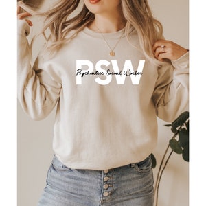 Peut inclure: Un sweat-shirt beige avec le texte "PSW Psychiatric Social Worker" imprimé en blanc.