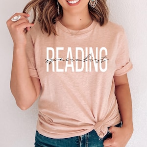 Puede incluir: Una camiseta rosa claro con el texto "READING specialist" en letras blancas y negras.