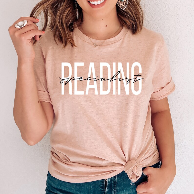 Puede incluir: Una camiseta rosa claro con el texto "READING specialist" en letras blancas y negras.