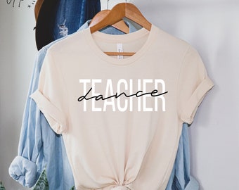 Op de afbeelding: Een crèmekleurig t-shirt met de woorden "TEACHER dance" in wit en zwart gedrukt. Het shirt hangt aan een houten hanger met een blauw overhemd en een hoed op de achtergrond.