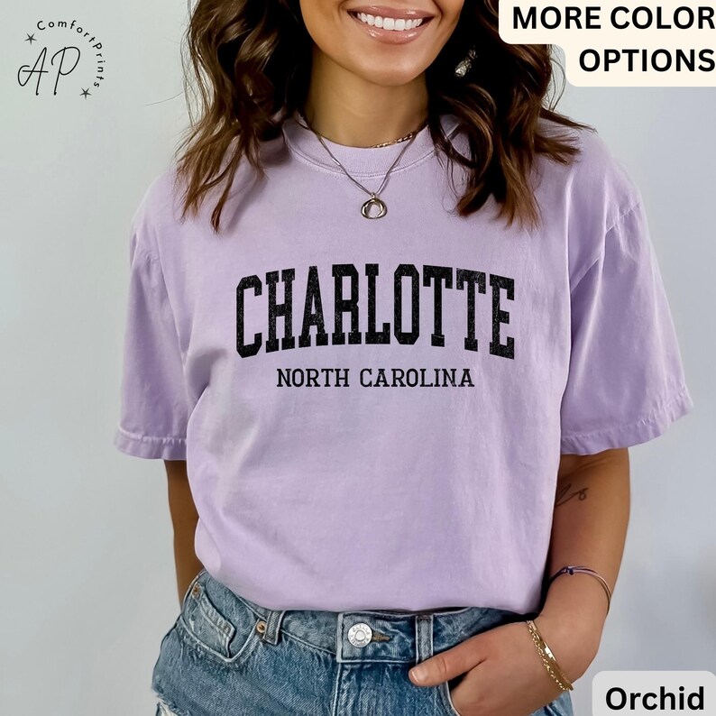Op de afbeelding: Een lichtpaars t-shirt met het woord "CHARLOTTE" in zwarte blokletters boven "NORTH CAROLINA". Het shirt heeft korte mouwen. Het shirt is effen van kleur en heeft korte mouwen.