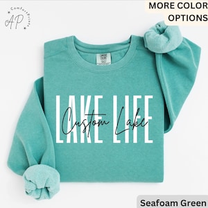 Könnte beinhalten: Meergrünes Sweatshirt mit den Wörtern "LAKE LIFE" in großen weißen Blockbuchstaben und "Custom Lake" in kursiver Schrift. Das Sweatshirt hat einen Rundhalsausschnitt und lange Ärmel.