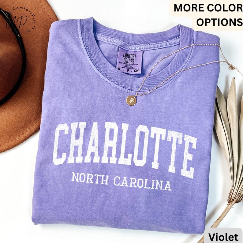Op de afbeelding: Een violetkleurig T-shirt met het woord "CHARLOTTE" in witte blokletters en "NORTH CAROLINA" eronder. Het shirt heeft een ronde hals en een klein label. Een gouden ketting met een hanger ligt op het shirt.