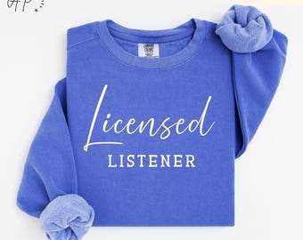 May include: A Flo Blue sweatshirt with the words "Licensed Listener" in white script. The long-sleeved shirt has a crew neck and the sleeves are cuffed. The Comfort Colors brand logo is visible on the tag.
