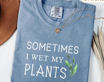 Peut inclure: T-shirt bleu jean avec le texte "SOMETIMES I WET MY PLANTS" en blanc. Le t-shirt a un col rond et des manches courtes. Un collier doré avec un pendentif est sur le t-shirt. Le t-shirt est plié sur une surface blanche.