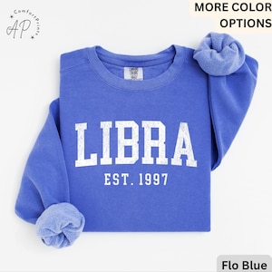 May include: A blue sweatshirt featuring the word "LIBRA" in white capital letters and "EST. 1997" below. The sleeves are rolled up. The sweatshirt is a solid blue colour.