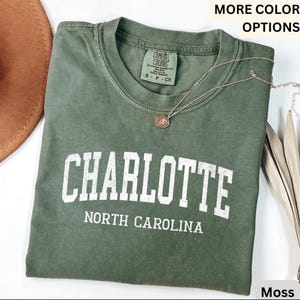 Op de afbeelding: Een mosgroen t-shirt met het woord "CHARLOTTE" in witte blokletters, met "NORTH CAROLINA" eronder. Een gouden ketting met een hanger ligt op het shirt. Het shirt is opgevouwen.