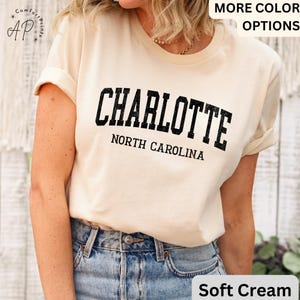 Op de afbeelding: Zacht cr&egrave;mekleurig T-shirt met het woord "CHARLOTTE" in vette, zwarte, gebogen letters en "NORTH CAROLINA" eronder. Het shirt heeft opgerolde mouwen en wordt gecombineerd met een spijkerbroek. De woorden "MORE COLOR OPTIONS" zijn zichtbaar.