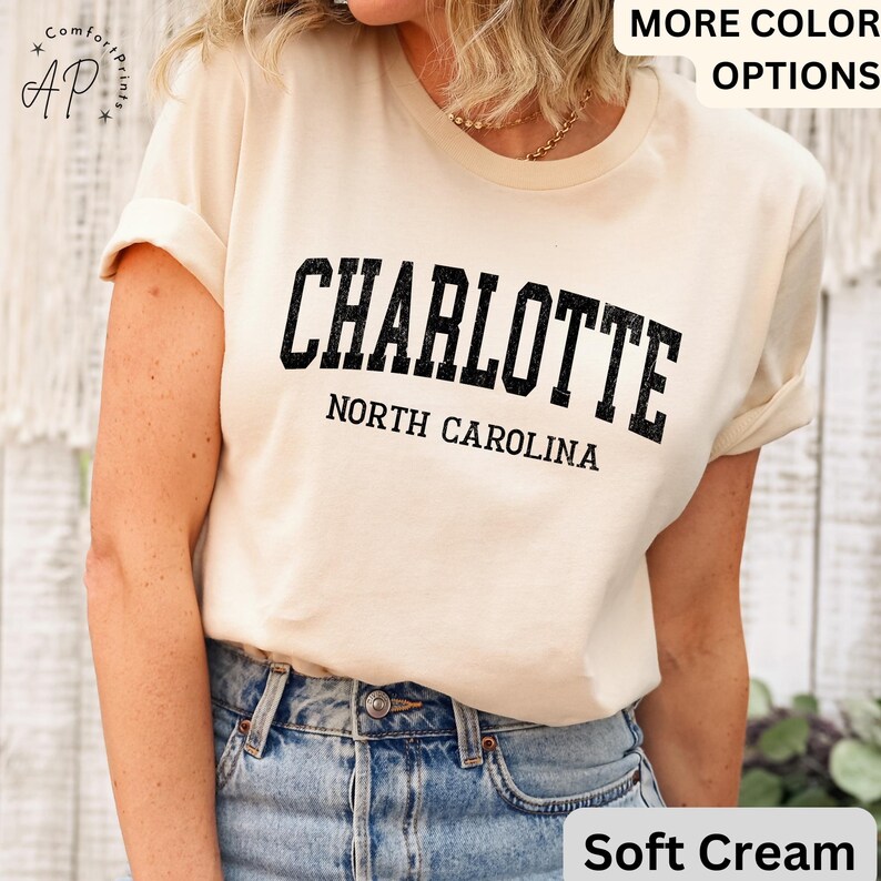 Op de afbeelding: Zacht cr&egrave;mekleurig T-shirt met het woord "CHARLOTTE" in vette, zwarte, gebogen letters en "NORTH CAROLINA" eronder. Het shirt heeft opgerolde mouwen en wordt gecombineerd met een spijkerbroek. De woorden "MORE COLOR OPTIONS" zijn zichtbaar.