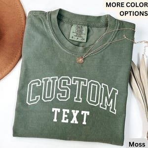 Puede incluir: Una camiseta verde musgo con el texto "CUSTOM TEXT" en blanco, impreso en una fuente de estilo universitario. La camiseta est&aacute; hecha de un material suave y c&oacute;modo.