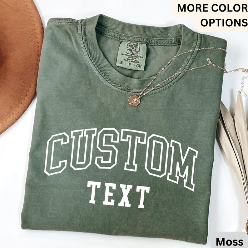 Puede incluir: Una camiseta verde musgo con el texto "CUSTOM TEXT" en blanco, impreso en una fuente de estilo universitario. La camiseta est&aacute; hecha de un material suave y c&oacute;modo.