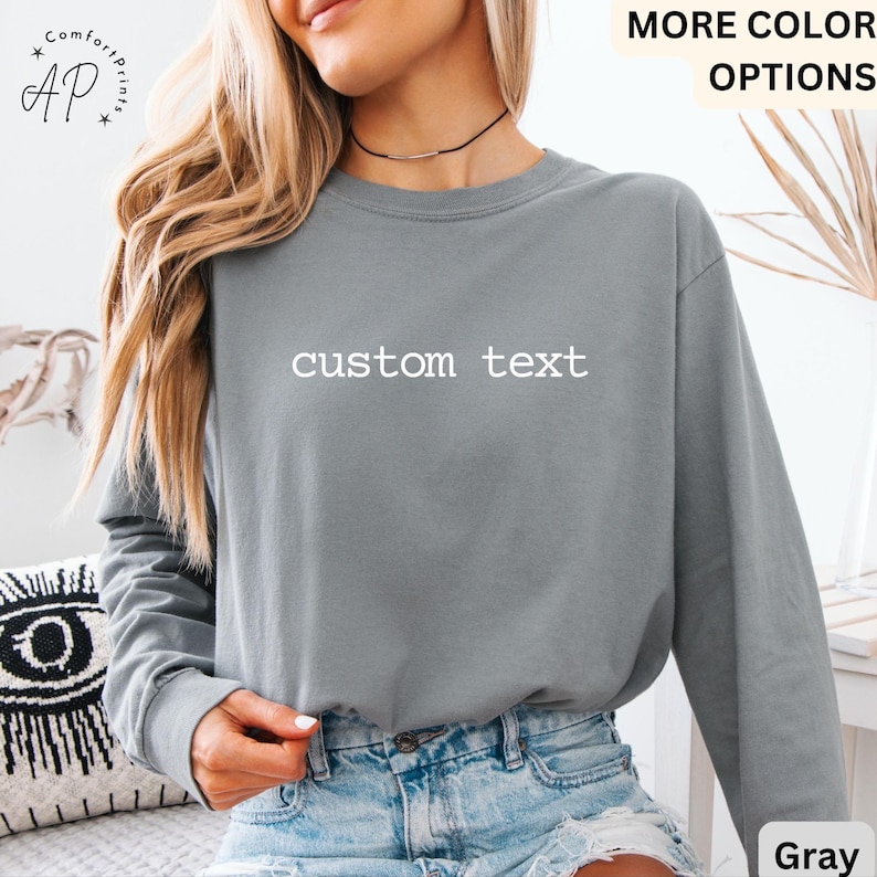 May include: Gray long-sleeve shirt with the text "custom text" in white. The shirt is a solid color and has a relaxed fit. The shirt is paired with blue denim shorts. The words "MORE COLOR OPTIONS" are visible.