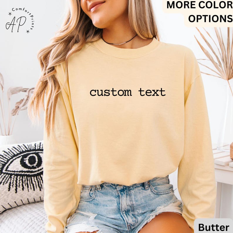 May include: A long-sleeved, butter-colored sweatshirt with the words "custom text" printed across the front. The sweatshirt is paired with denim shorts. The image also includes the text "MORE COLOR OPTIONS" and the brand logo "Comfort Prints".