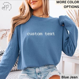 May include: A long-sleeved, blue jean-colored shirt with the words "custom text" printed in white across the chest. The shirt has a crew neck and long sleeves. The model is wearing blue jean shorts. The shirt is part of the "More Color Options" collection.