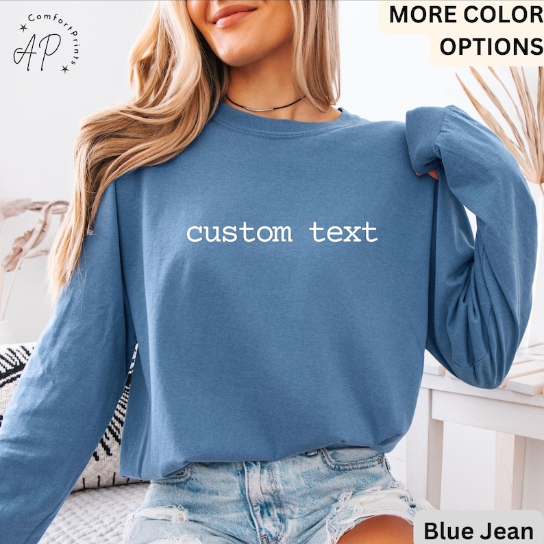 May include: A long-sleeved, blue jean-colored shirt with the words "custom text" printed in white across the chest. The shirt has a crew neck and long sleeves. The model is wearing blue jean shorts. The shirt is part of the "More Color Options" collection.