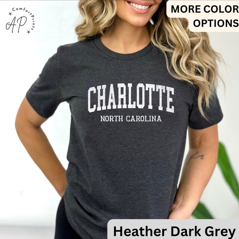 Op de afbeelding: Donkergrijs gem&ecirc;leerd T-shirt met het woord "CHARLOTTE" in witte hoofdletters boven "NORTH CAROLINA". Het shirt heeft korte mouwen en een ronde hals. De tekst "MORE COLOR OPTIONS" is zichtbaar.