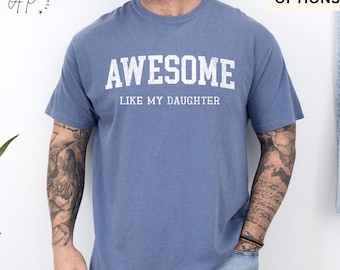 May include: Blue jean colored t-shirt with the word "AWESOME" in white block letters above the phrase "LIKE MY DAUGHTER". The shirt is a short-sleeved crew neck.