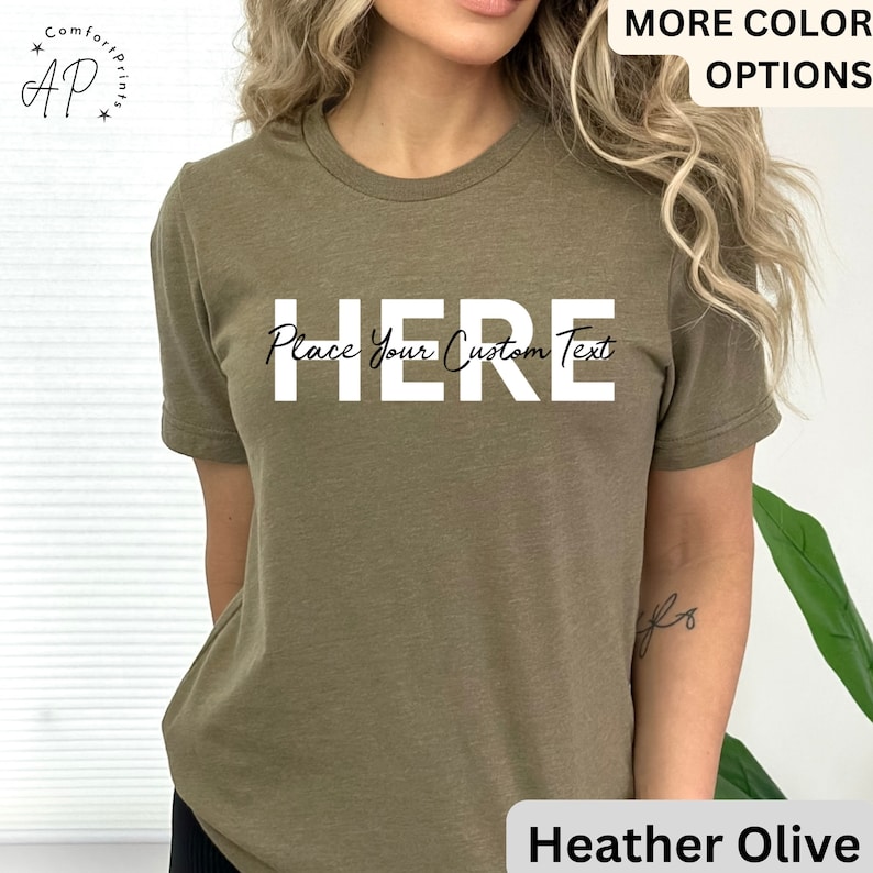 Puede incluir: Camiseta verde oliva jaspeada con la palabra "HERE" en letras blancas grandes. Debajo de la palabra "HERE" est&aacute; el texto "Place Your Custom Text". La camiseta tiene mangas cortas y cuello redondo.