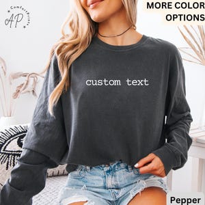 May include: A dark gray long-sleeve shirt with the text "custom text" in white. The shirt is worn with denim shorts. The words "MORE COLOR OPTIONS" are visible in the upper right corner. The shirt is part of the Comfort Prints collection.