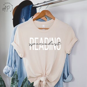 Puede incluir: Una camiseta beige con el texto "READING specialist" impreso en blanco y negro en la parte delantera.