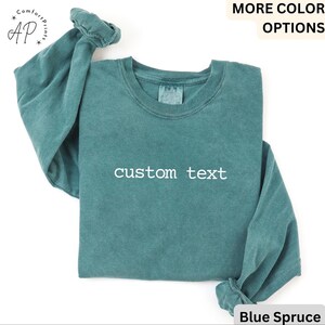 May include: A teal-colored long-sleeve sweatshirt with the words "custom text" printed in white across the front. The sweatshirt has a crew neck and the sleeves are cuffed. The words "MORE COLOR OPTIONS" are at the top, and "Blue Spruce" is at the bottom.