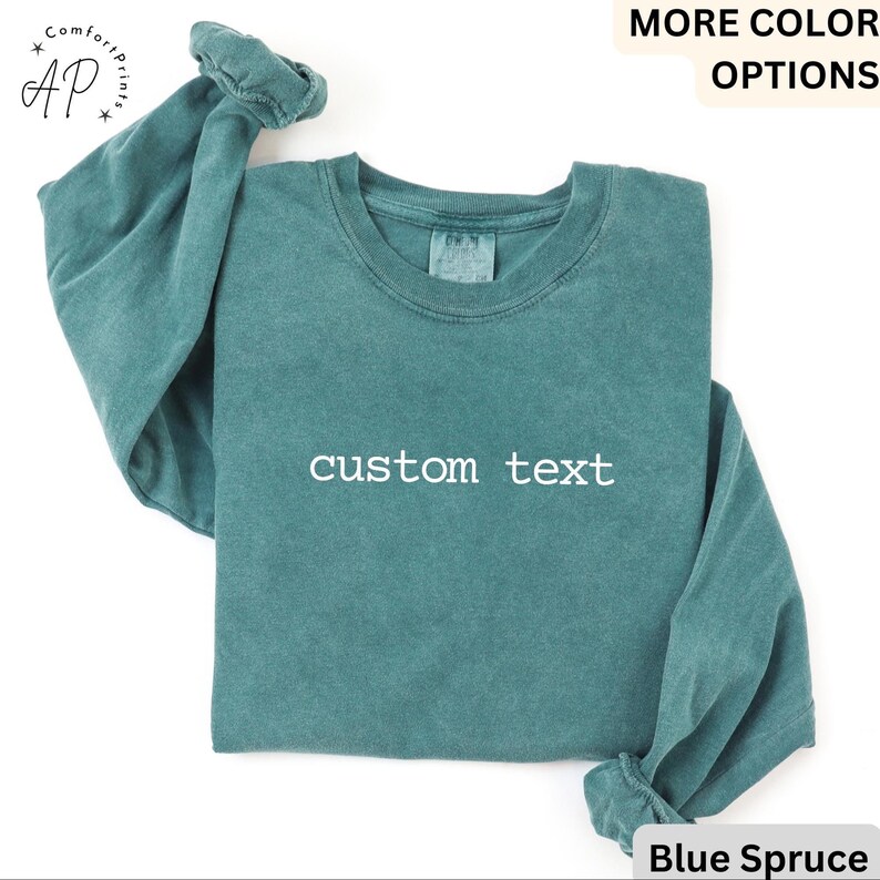 May include: A teal-colored long-sleeve sweatshirt with the words "custom text" printed in white across the front. The sweatshirt has a crew neck and the sleeves are cuffed. The words "MORE COLOR OPTIONS" are at the top, and "Blue Spruce" is at the bottom.