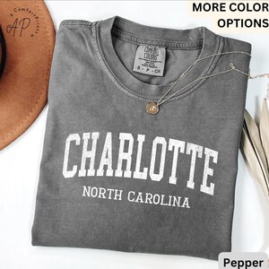 Op de afbeelding: Een grijs t-shirt met het woord "CHARLOTTE" in grote witte letters en "NORTH CAROLINA" eronder. Het shirt heeft een ronde hals en een klein label. Een zilveren ketting met een hanger ligt over het shirt.