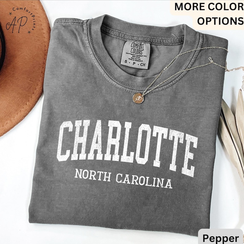 Op de afbeelding: Een grijs t-shirt met het woord "CHARLOTTE" in grote witte letters en "NORTH CAROLINA" eronder. Het shirt heeft een ronde hals en een klein label. Een zilveren ketting met een hanger ligt over het shirt.