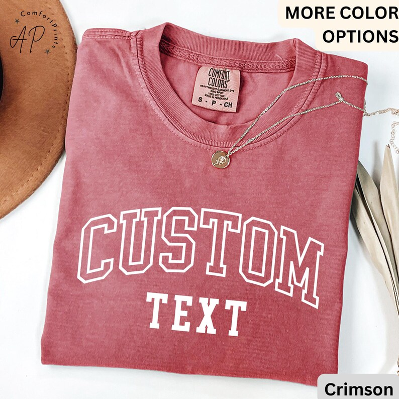 Puede incluir: Camiseta roja carmes&iacute; con texto blanco que dice "CUSTOM TEXT" en una fuente universitaria.