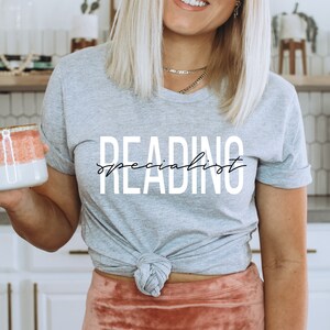 Puede incluir: Una camiseta gris con el texto "READING specialist" en letras blancas con una fuente de letra cursiva negra.