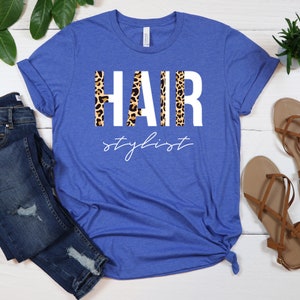 May include: A royal blue t-shirt with the word "HAIR" in white letters with a leopard print pattern behind the letters. The text "stylist" is written in white cursive below the word "HAIR".