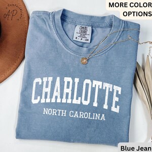 Op de afbeelding: Een blauw spijkerbroekkleurig t-shirt met het woord "CHARLOTTE" in grote witte blokletters. Daaronder staat de tekst "NORTH CAROLINA" gedrukt. De shirt heeft een ronde hals en korte mouwen. Een gouden ketting met een hanger ligt over het shirt.
