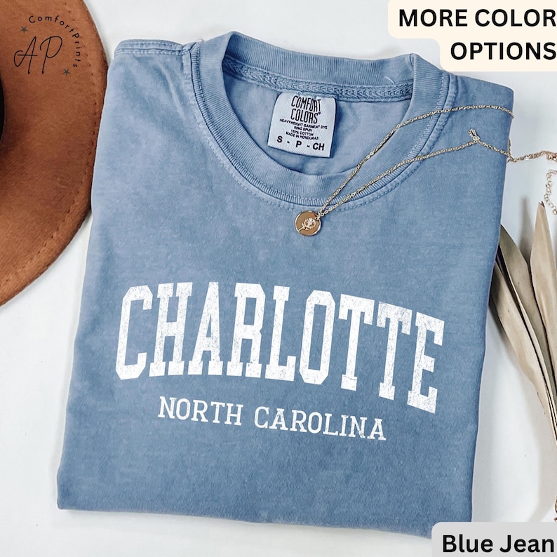 Op de afbeelding: Een blauw spijkerbroekkleurig t-shirt met het woord "CHARLOTTE" in grote witte blokletters. Daaronder staat de tekst "NORTH CAROLINA" gedrukt. De shirt heeft een ronde hals en korte mouwen. Een gouden ketting met een hanger ligt over het shirt.