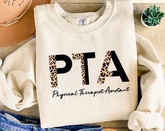 Op de afbeelding: Een crèmekleurig sweatshirt met de letters "PTA" in zwart op een luipaardprint achtergrond. De tekst "Physical Therapist Assistant" is onder de letters gedrukt.
