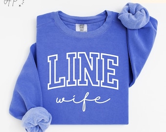 May include: A Flo Blue sweatshirt featuring the text "LINE wife" in white, block lettering and script. The crew neck sweatshirt has long sleeves with cuffed wrists. The words "MORE COLOR OPTIONS" are visible in the top right corner.