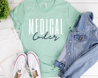 May include: A light green t-shirt with the text "MEDICAL Coder" in white and dark blue lettering. The shirt is laid flat on a white surface with a pair of white sneakers, a white pair of jeans, and a light blue denim jacket.