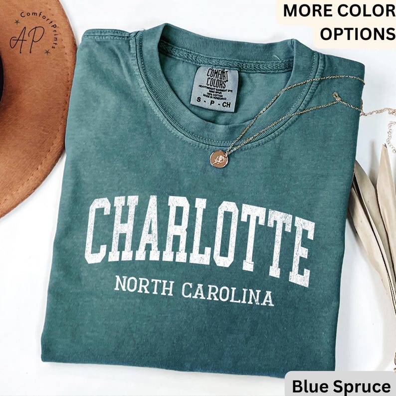 Op de afbeelding: Een blauwgroen t-shirt met het woord "CHARLOTTE" in witte blokletters boven "NORTH CAROLINA". Het shirt heeft een ronde hals en korte mouwen. Een gouden ketting met een hanger is over het shirt gedrapeerd.
