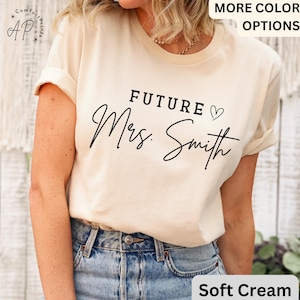 May include: Soft cream-colored t-shirt with the text "FUTURE Mrs. Smith" in black script. The shirt has rolled-up sleeves and is paired with blue jeans. The top also has the words "MORE COLOR OPTIONS" in the top right corner.