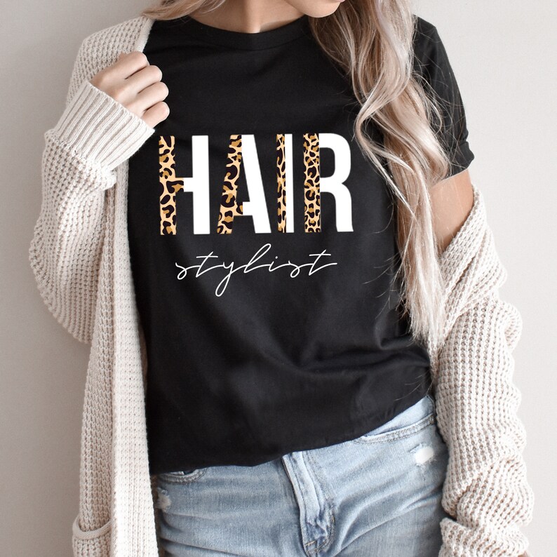 May include: Black t-shirt with the word "HAIR" in white letters with a leopard print pattern behind the letters. The text "stylist" is written in white cursive below the word "HAIR".